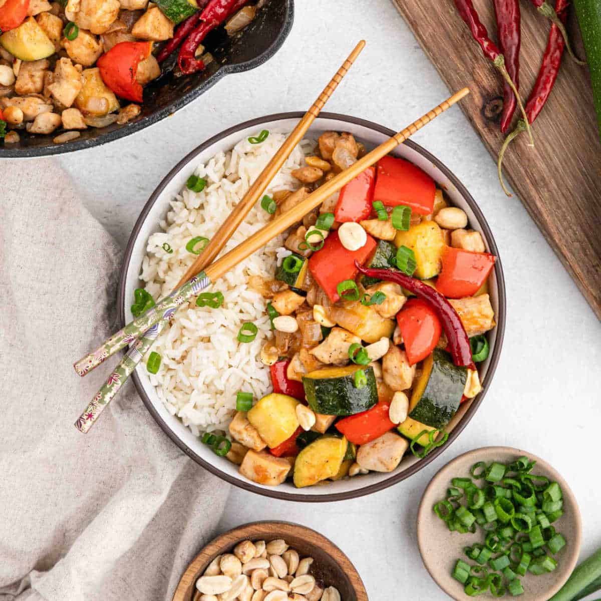 Kung Pao Chicken served in a white bowl with chopsticks, garnished with green onions and crushed peanuts, showing colorful vegetables and tender chicken pieces