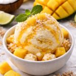 Mango coconut nice cream vegan served in a white bowl with fresh mango slices and toasted coconut flakes on top, photographed on a bright tropical-inspired table setting