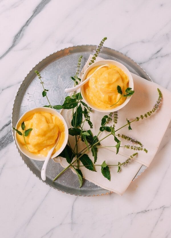 Overhead flat lay of ingredients for mango coconut nice cream — frozen mango chunks in a bowl, a can of full-fat coconut milk, fresh lime, sea salt, and fresh ginger on a light marble surface