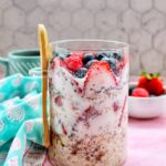 Overhead shot of two glass mason jars filled with creamy overnight oats, topped with fresh strawberries, blueberries, raspberries, and blackberries, set on a white linen cloth with a wooden spoon and scattered berries around them in warm morning light