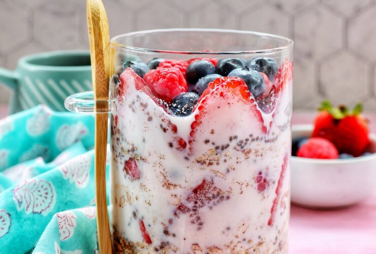 Overhead shot of two glass mason jars filled with creamy overnight oats, topped with fresh strawberries, blueberries, raspberries, and blackberries, set on a white linen cloth with a wooden spoon and scattered berries around them in warm morning light