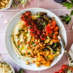 Overhead shot of a beautifully plated roasted chickpea hummus bowl with golden crispy chickpeas, rainbow vegetables, and creamy hummus on a white ceramic bowl with a linen napkin and fresh herbs