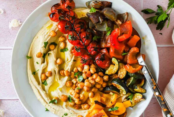 Overhead shot of a beautifully plated roasted chickpea hummus bowl with golden crispy chickpeas, rainbow vegetables, and creamy hummus on a white ceramic bowl with a linen napkin and fresh herbs