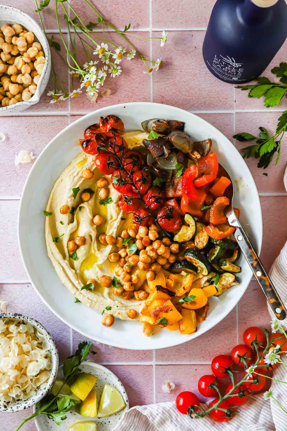Overhead shot of a beautifully plated roasted chickpea hummus bowl with golden crispy chickpeas, rainbow vegetables, and creamy hummus on a white ceramic bowl with a linen napkin and fresh herbs