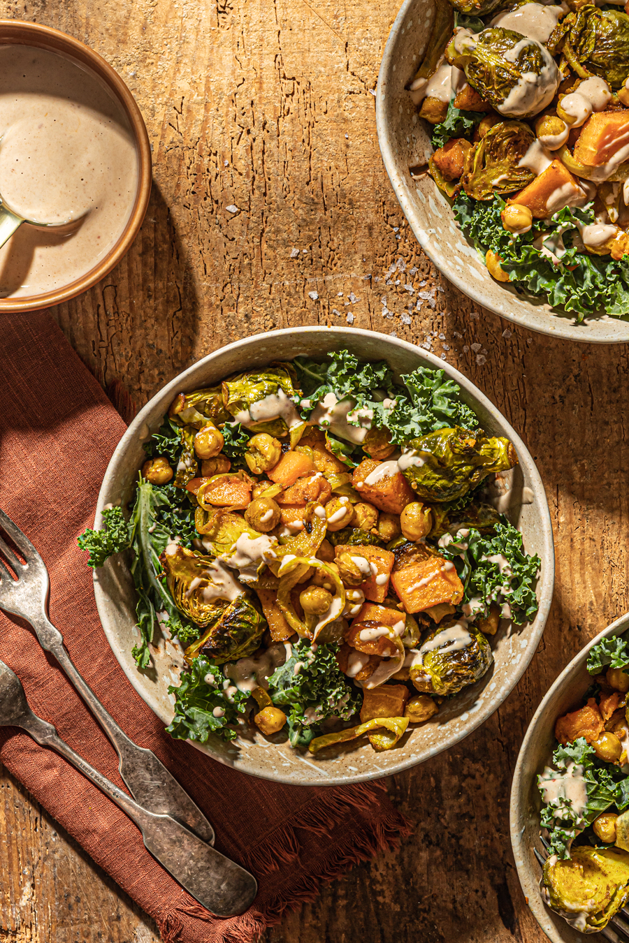 Flat lay of roasted chickpea hummus bowl ingredients including chickpeas on a baking sheet, tahini, fresh vegetables, lemons, and spices arranged on a wooden cutting board