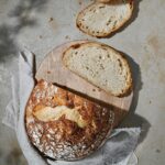 Golden brown sourdough bread loaf with perfectly crispy crust and airy crumb, artfully placed on rustic wooden cutting board with wheat stalks