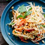 Overhead shot of a beautifully plated healthy Thai papaya salad som tam in a wide shallow bowl, garnished with crushed peanuts, fresh lime wedges, Thai basil, and sliced red chilies on a wooden table with tropical styling