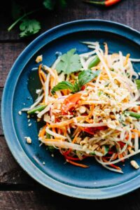 Overhead shot of a beautifully plated healthy Thai papaya salad som tam in a wide shallow bowl, garnished with crushed peanuts, fresh lime wedges, Thai basil, and sliced red chilies on a wooden table with tropical styling