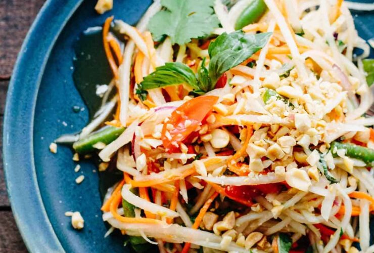 Overhead shot of a beautifully plated healthy Thai papaya salad som tam in a wide shallow bowl, garnished with crushed peanuts, fresh lime wedges, Thai basil, and sliced red chilies on a wooden table with tropical styling