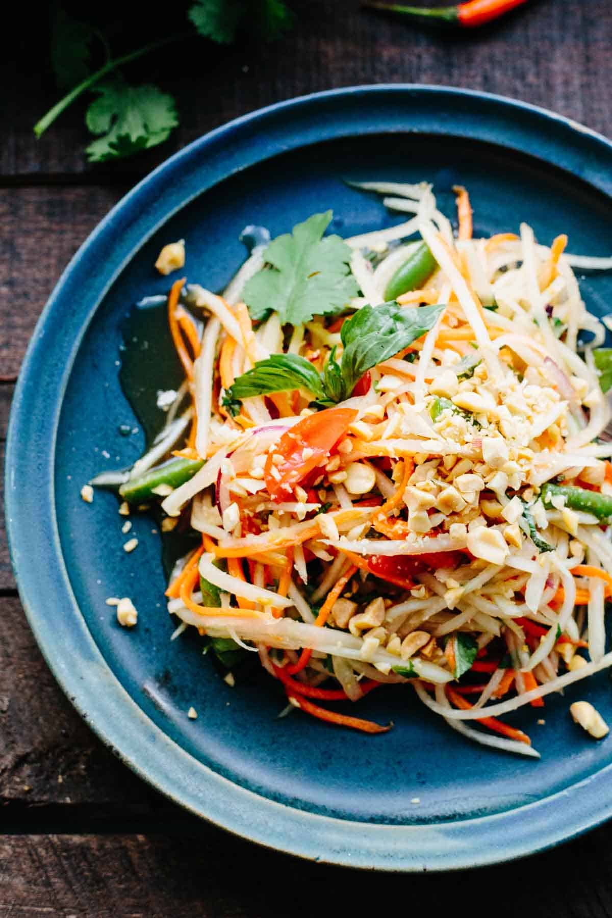Overhead shot of a beautifully plated healthy Thai papaya salad som tam in a wide shallow bowl, garnished with crushed peanuts, fresh lime wedges, Thai basil, and sliced red chilies on a wooden table with tropical styling