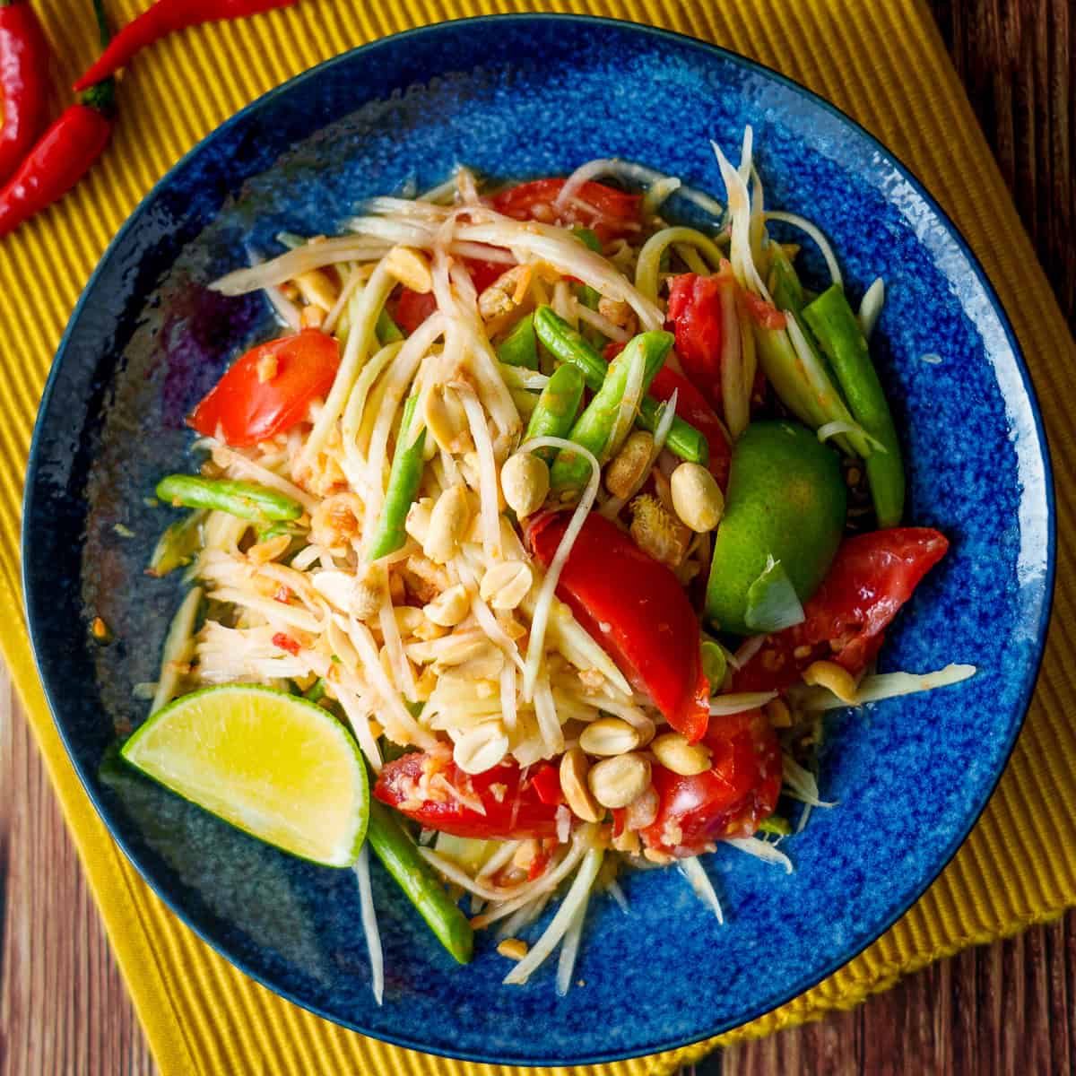 Close-up serving shot of healthy Thai papaya salad som tam in a rustic bowl with chopsticks resting alongside, highlighting the golden crushed peanuts, bright red chili slices, and glistening lime dressing coating the fine papaya shreds