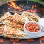 Golden crispy campfire quesadillas served on a rustic wooden board outdoors near a campfire, cut into wedges with melted cheese pulling apart