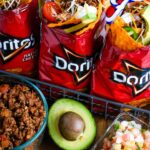 Campfire walking tacos served in open Dorito bags with seasoned beef, shredded cheese, sour cream, salsa, and fresh toppings on a rustic wooden camp table beside a glowing campfire at dusk