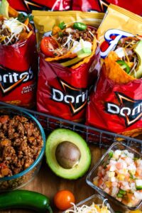 Campfire walking tacos served in open Dorito bags with seasoned beef, shredded cheese, sour cream, salsa, and fresh toppings on a rustic wooden camp table beside a glowing campfire at dusk