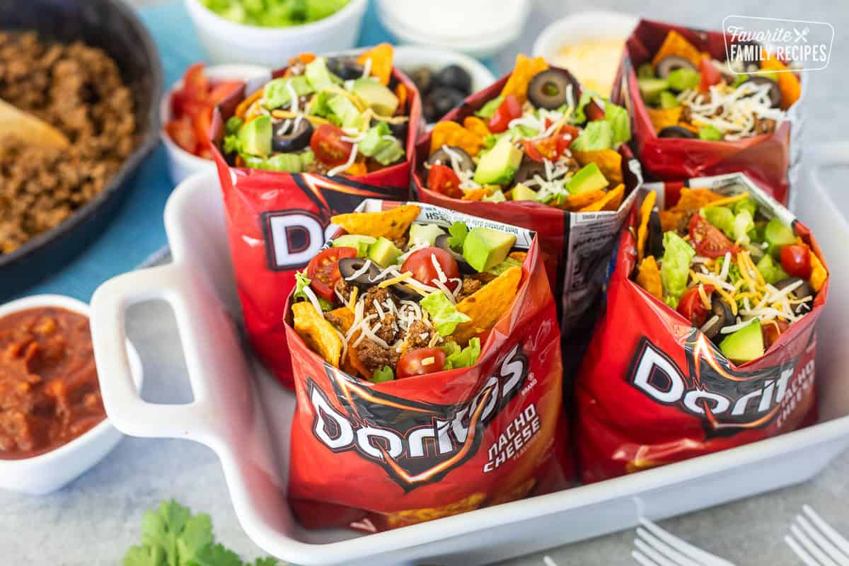 Campfire Walking Tacos — Easy Dorito Bag Tacos with Seasoned Beef & All the Toppings 28 Close-up overhead shot of an open Dorito bag filled with seasoned beef, melted cheese, fresh pico de gallo, sliced avocado, and a drizzle of sour cream, with a lime wedge on the side and blurred campfire in the background