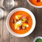 A rustic white ceramic bowl filled with vibrant red chilled gazpacho garnished with diced cucumber, cherry tomatoes, a drizzle of olive oil, and fresh basil leaves, photographed on a linen napkin in bright natural light