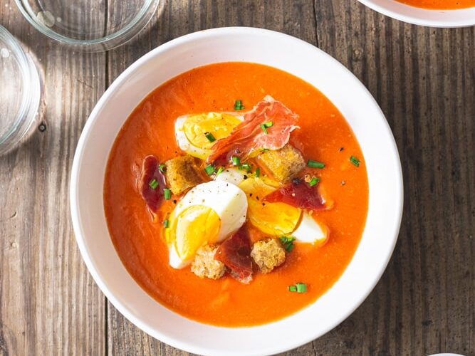 A rustic white ceramic bowl filled with vibrant red chilled gazpacho garnished with diced cucumber, cherry tomatoes, a drizzle of olive oil, and fresh basil leaves, photographed on a linen napkin in bright natural light