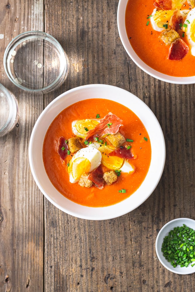 A rustic white ceramic bowl filled with vibrant red chilled gazpacho garnished with diced cucumber, cherry tomatoes, a drizzle of olive oil, and fresh basil leaves, photographed on a linen napkin in bright natural light