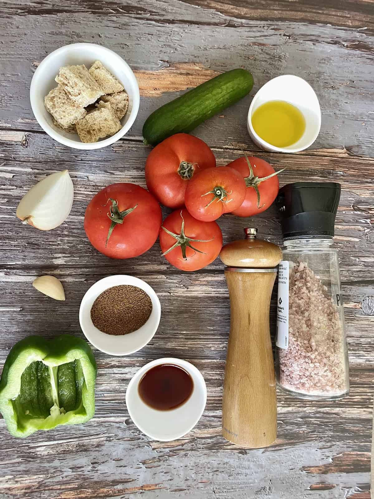 A flat-lay overhead shot of chilled gazpacho ingredients arranged on a white marble surface — ripe red tomatoes, cucumber, red bell pepper, garlic cloves, a small pitcher of olive oil, and a bottle of sherry vinegar