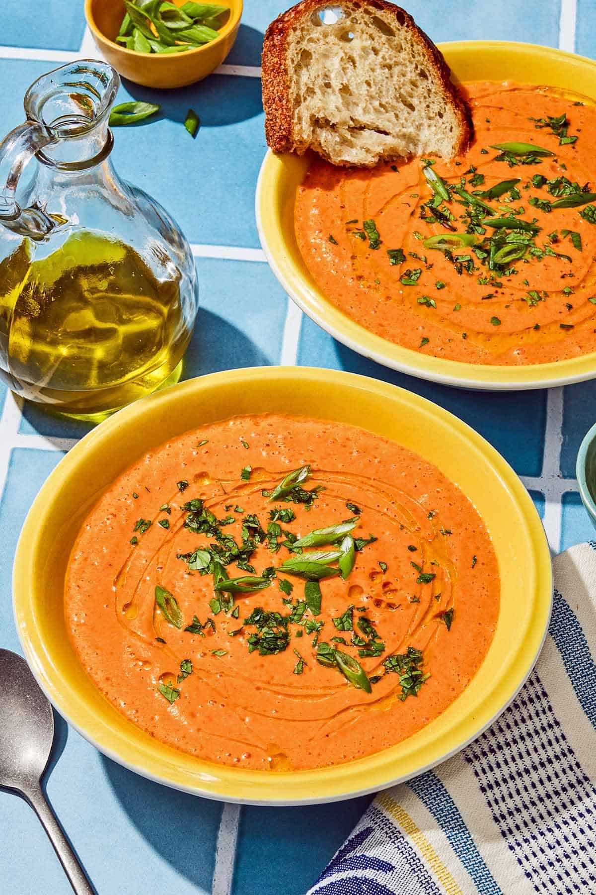 A close-up shot of a chilled gazpacho bowl showing the smooth crimson soup with vibrant garnishes of finely diced cucumber, red onion, and a swirl of golden olive oil, with a rustic wooden spoon resting on the rim