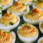 Classic deviled eggs arranged on a white ceramic serving platter, topped with smoked paprika and fresh chives, shot from above on a rustic wooden picnic table