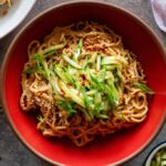Cold sesame noodles in a wide bowl topped with sliced cucumbers, shredded carrots, chopped scallions, sesame seeds, and a drizzle of chili oil, photographed overhead on a light linen surface