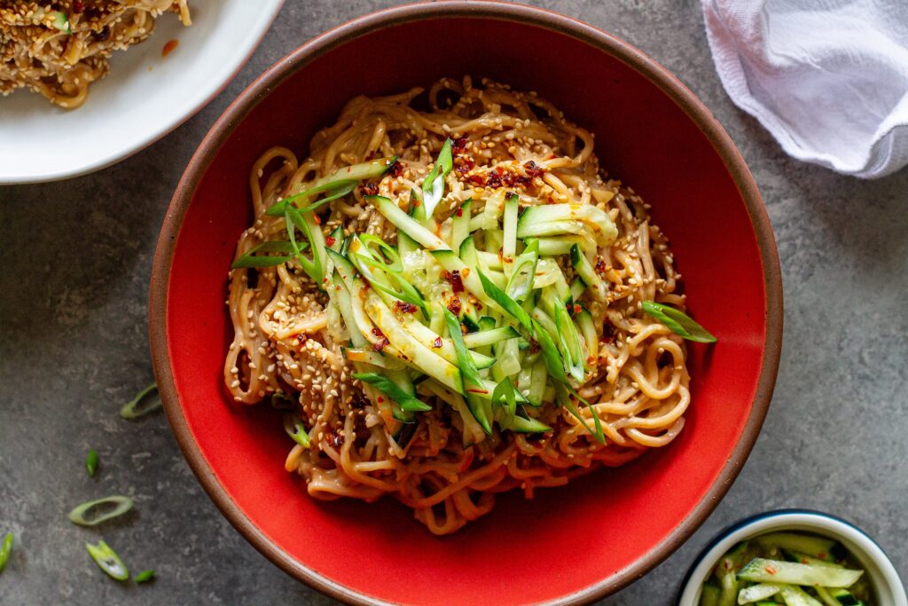 Cold Sesame Noodles — No-Cook Spicy Peanut Noodles Ready in 15 Minutes 26 Cold sesame noodles in a wide bowl topped with sliced cucumbers, shredded carrots, chopped scallions, sesame seeds, and a drizzle of chili oil, photographed overhead on a light linen surface
