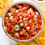 A rustic bowl of vibrant fresh pico de gallo with diced tomatoes, white onion, green jalapeño, and cilantro, shot overhead on a wooden board with tortilla chips alongside