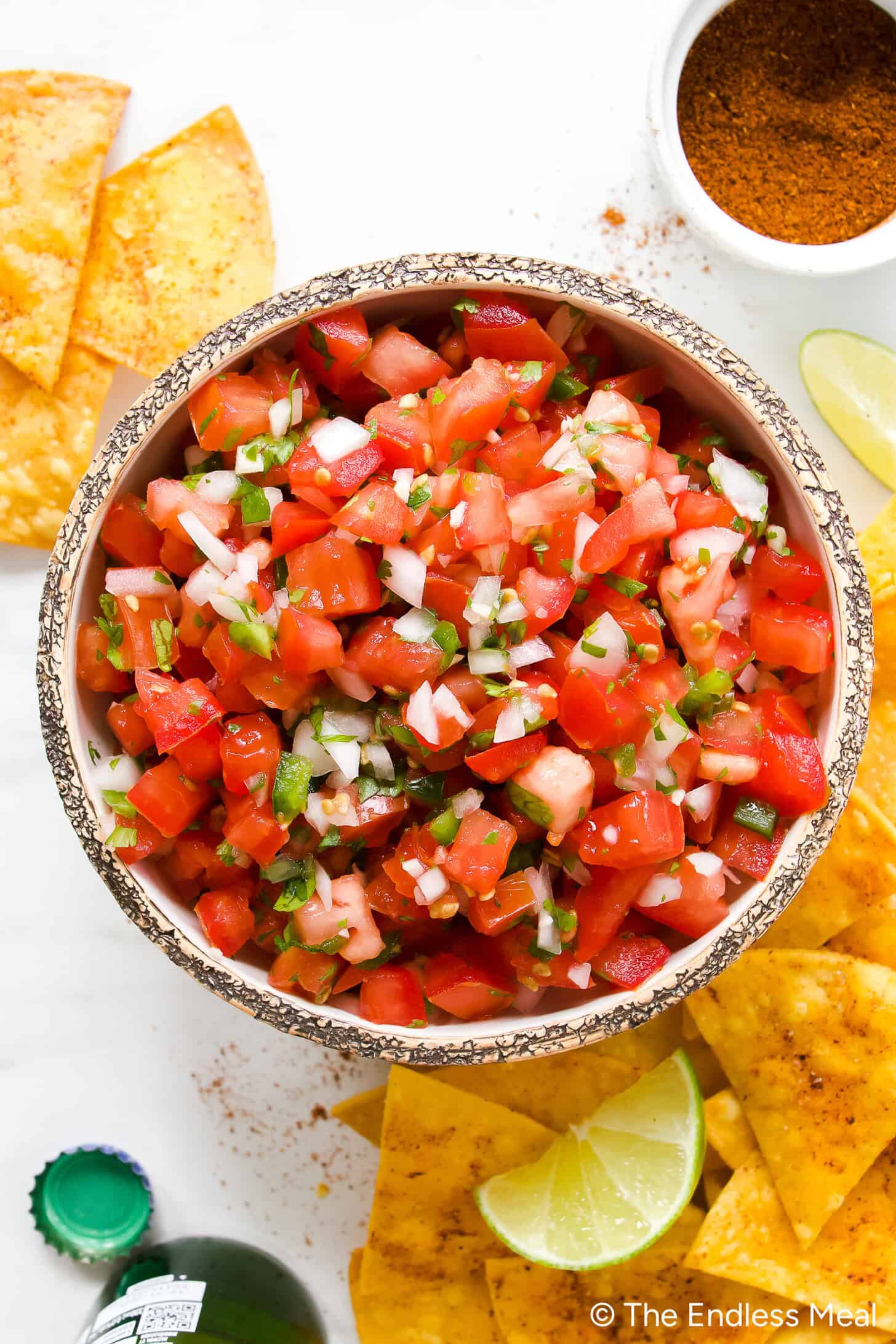 A rustic bowl of vibrant fresh pico de gallo with diced tomatoes, white onion, green jalapeño, and cilantro, shot overhead on a wooden board with tortilla chips alongside