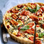 Grilled vegetable and goat cheese tart on a rustic wooden board, golden whole wheat crust, vibrant zucchini, bell peppers, and eggplant topped with creamy white goat cheese and fresh herbs