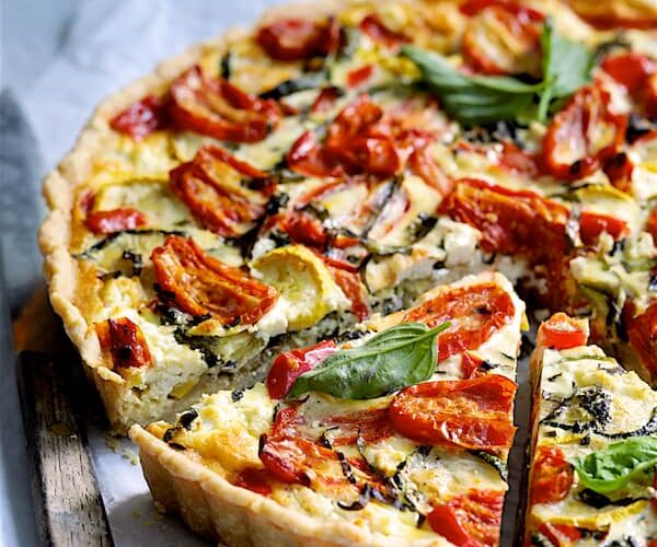 Grilled vegetable and goat cheese tart on a rustic wooden board, golden whole wheat crust, vibrant zucchini, bell peppers, and eggplant topped with creamy white goat cheese and fresh herbs