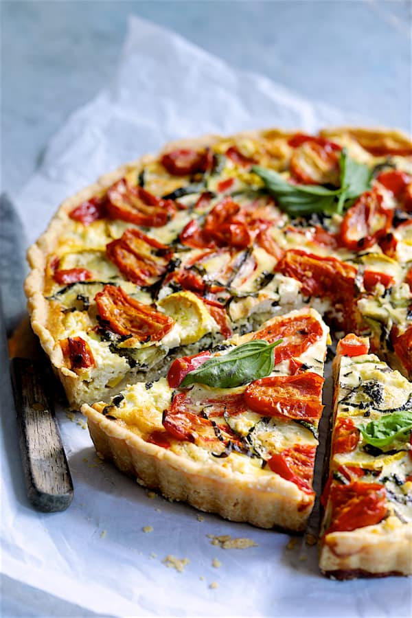 Grilled vegetable and goat cheese tart on a rustic wooden board, golden whole wheat crust, vibrant zucchini, bell peppers, and eggplant topped with creamy white goat cheese and fresh herbs