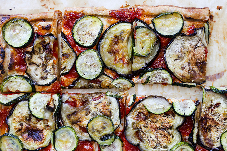 Step-by-step ingredients flat lay showing sliced zucchini, eggplant, bell peppers, red onion, crumbled goat cheese, fresh thyme, olive oil, and whole wheat pastry dough on a marble surface