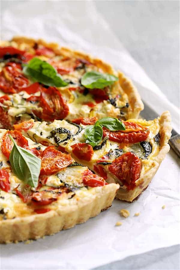 Close-up of a slice of grilled vegetable and goat cheese tart on a white plate, showing flaky whole wheat crust layers, colorful grilled vegetables, and melted goat cheese with fresh thyme garnish