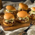 Instant pot pulled pork for camping served on slider buns with coleslaw on a wooden picnic table outdoors, golden hour light