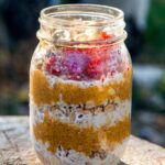 A rustic enamel camping mug filled with creamy overnight camping oatmeal topped with dried cranberries, chopped walnuts, and a drizzle of honey, set on a wooden picnic table with a campfire blurred in the background