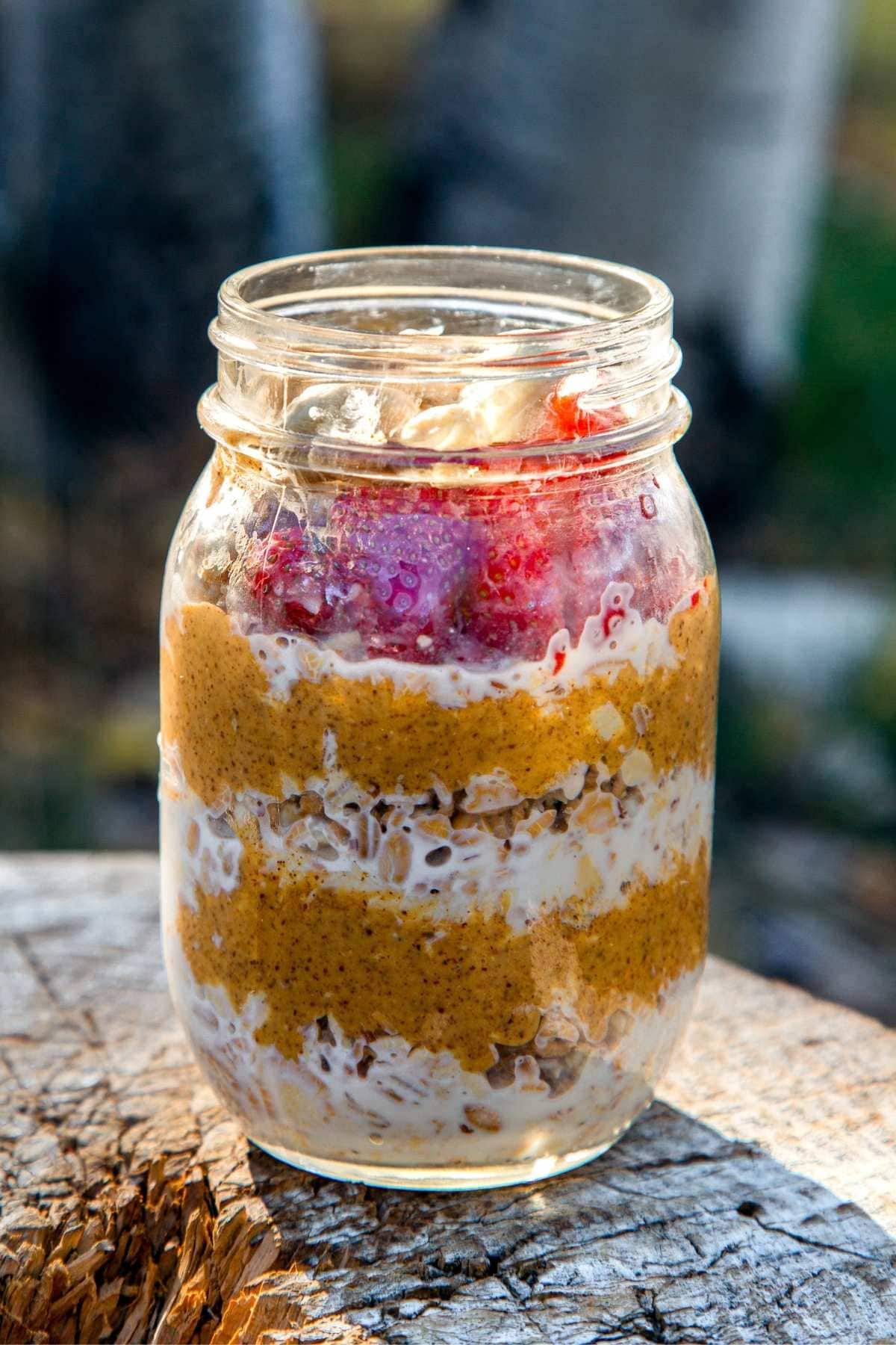 Overnight Camping Oatmeal — The Easy, Nourishing Breakfast You Prep Before You Even Leave Home 26 A rustic enamel camping mug filled with creamy overnight camping oatmeal topped with dried cranberries, chopped walnuts, and a drizzle of honey, set on a wooden picnic table with a campfire blurred in the background