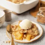 Golden peach cobbler in a white baking dish with biscuit topping, served with a scoop of vanilla ice cream melting over the top, photographed on a rustic wooden table with fresh peaches alongside