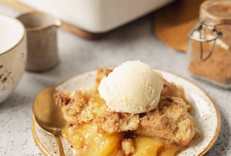 Golden peach cobbler in a white baking dish with biscuit topping, served with a scoop of vanilla ice cream melting over the top, photographed on a rustic wooden table with fresh peaches alongside