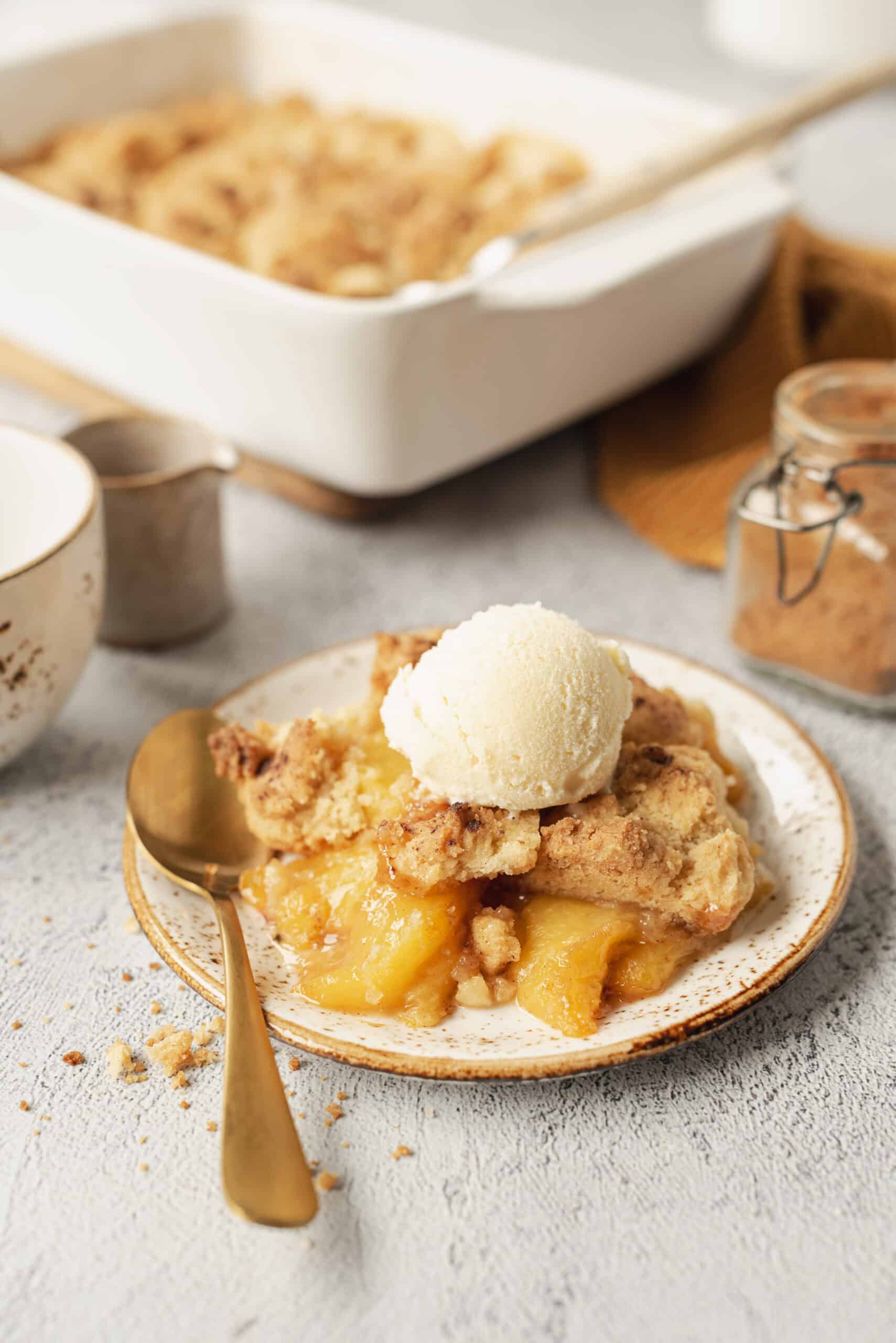 Golden peach cobbler in a white baking dish with biscuit topping, served with a scoop of vanilla ice cream melting over the top, photographed on a rustic wooden table with fresh peaches alongside