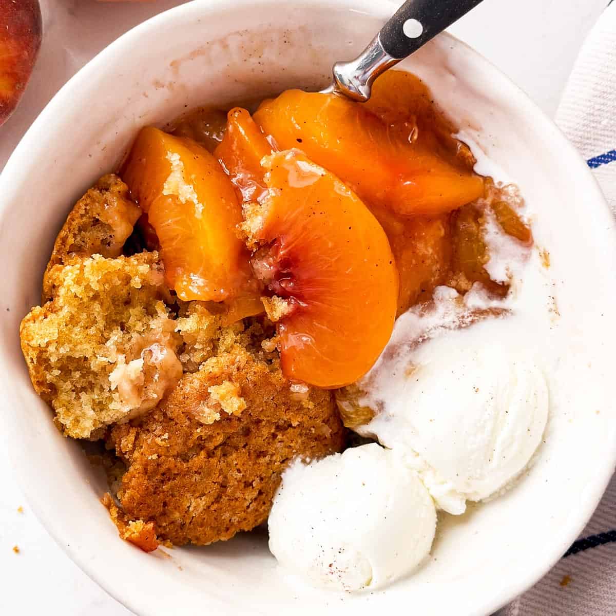 Overhead flat lay of peach cobbler ingredients including sliced fresh peaches, flour, cold cubed butter, brown sugar, cinnamon, and baking powder on a marble surface