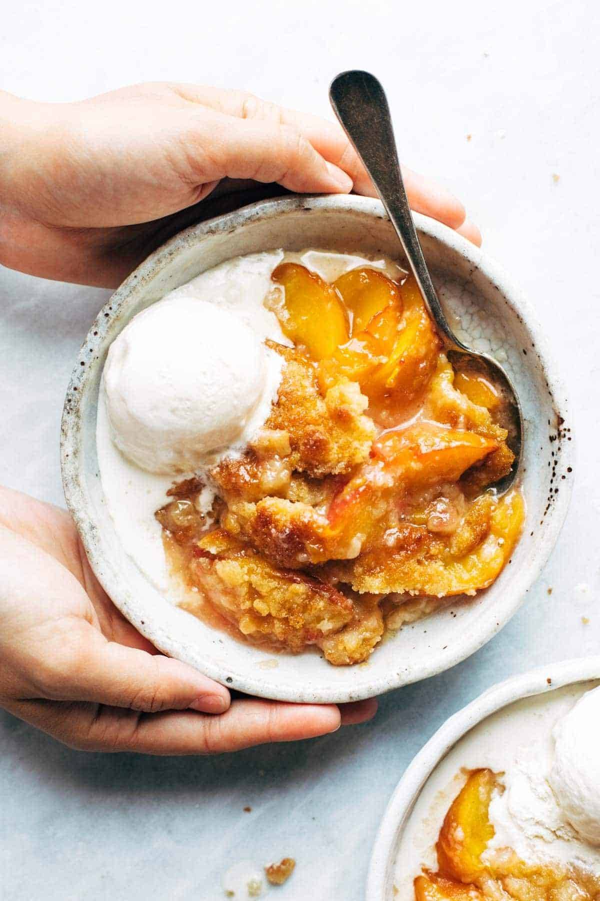 Close-up of a spoonful of peach cobbler being lifted from the dish, showing glossy golden peach filling and flaky biscuit topping, with melting vanilla ice cream in soft focus background