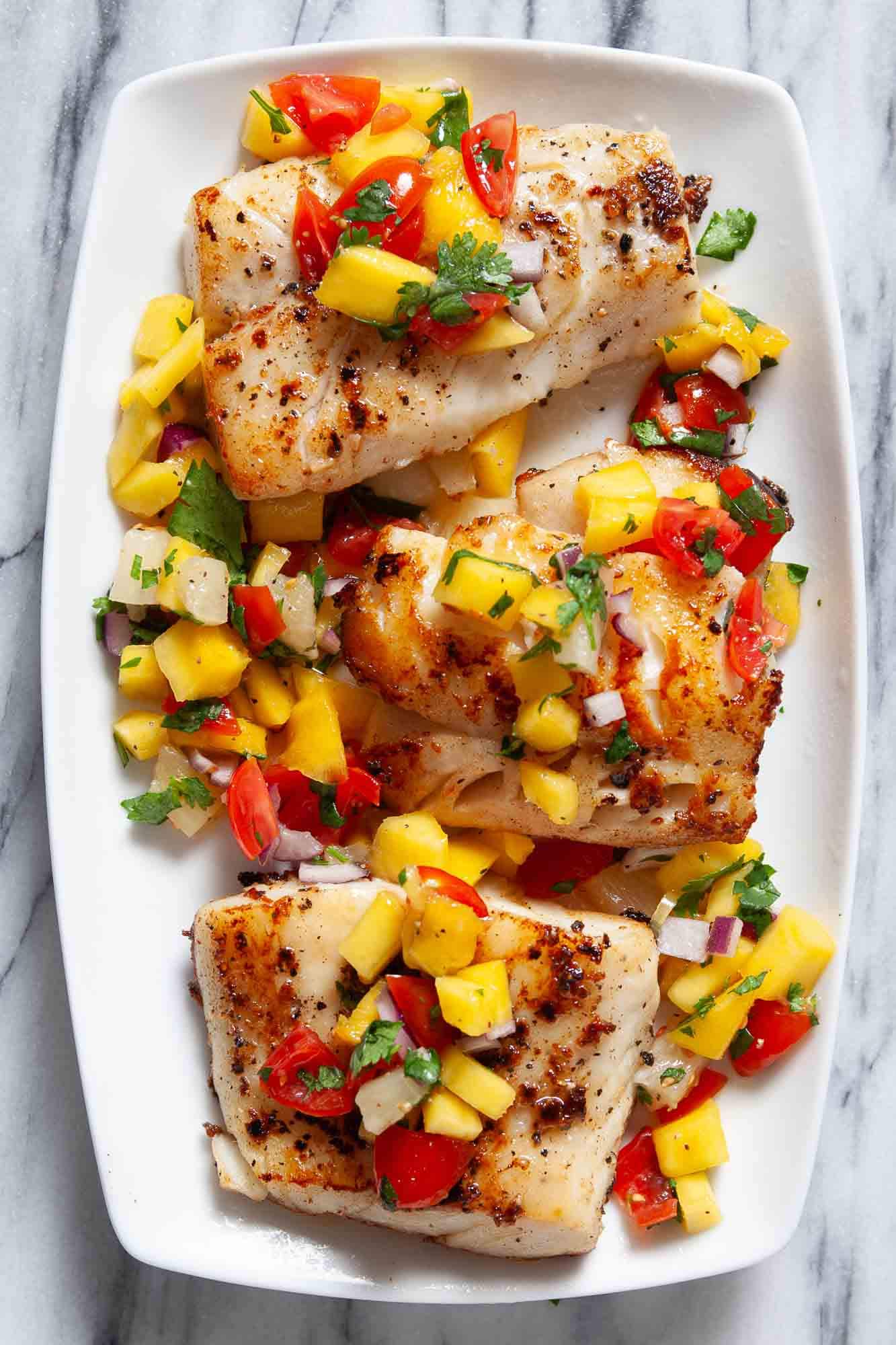 Close-up overhead shot of peach mango salsa spooned generously over a grilled mahi-mahi fillet on a plate, with lime wedges and extra cilantro scattered around