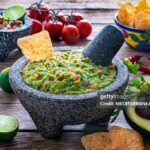 restaurant style guacamole served in a stone molcajete bowl with tortilla chips, fresh lime wedges, and cilantro garnish on a rustic wooden board