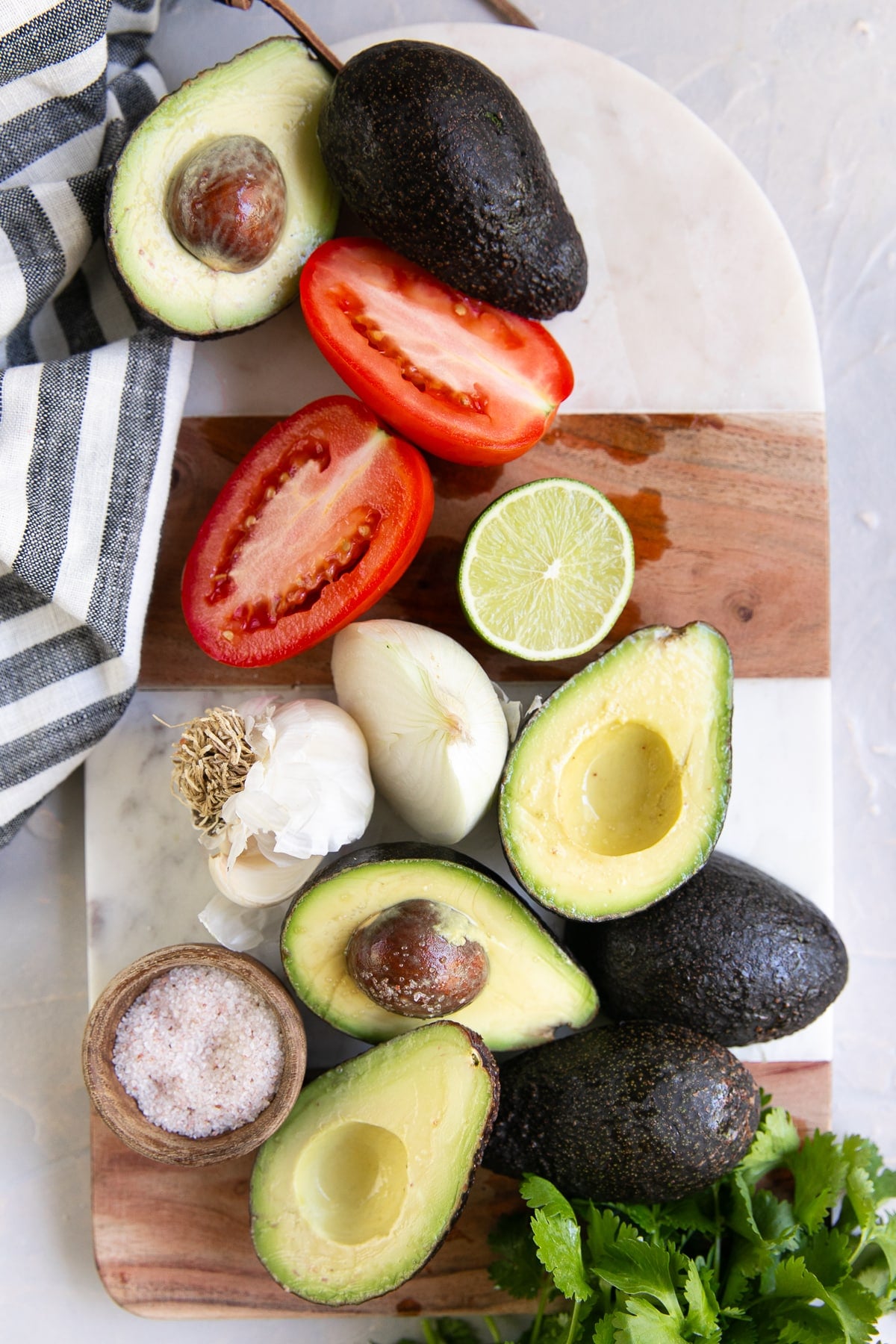 overhead flat lay of guacamole ingredients including ripe hass avocados cut in half, fresh limes, white onion, cilantro, serrano peppers, and sea salt on a white marble surface