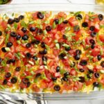Seven Layer Dip beautifully plated in a wide glass dish with colorful layers of beans, sour cream, guacamole, cheese, tomatoes, olives, and green onions served with tortilla chips