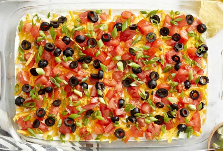 Seven Layer Dip beautifully plated in a wide glass dish with colorful layers of beans, sour cream, guacamole, cheese, tomatoes, olives, and green onions served with tortilla chips