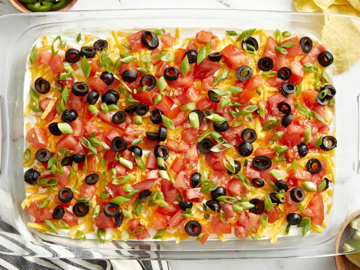 Seven Layer Dip beautifully plated in a wide glass dish with colorful layers of beans, sour cream, guacamole, cheese, tomatoes, olives, and green onions served with tortilla chips