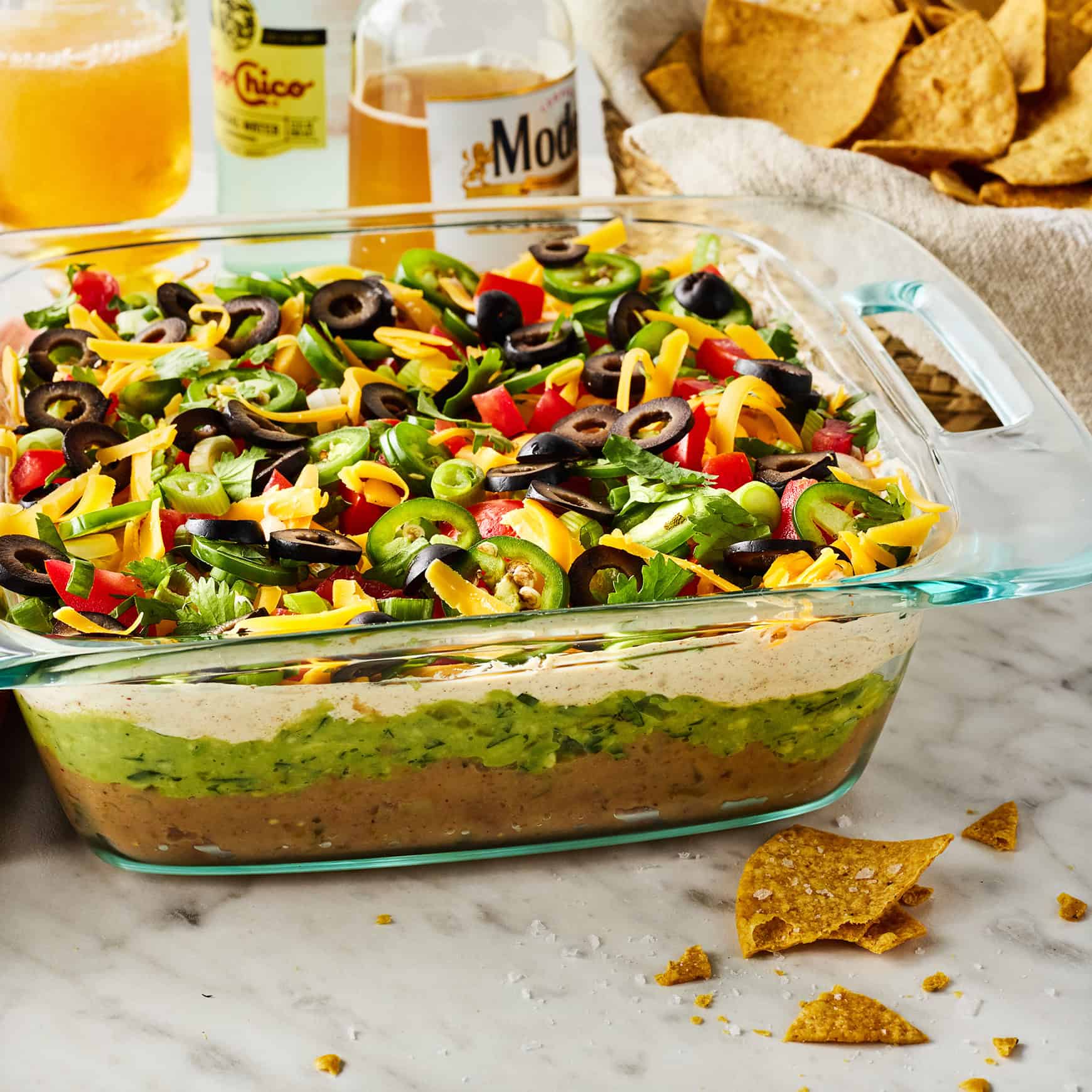 Seven Layer Dip ingredients laid out flat lay overhead including avocados, refried beans, sour cream, cheddar cheese, tomatoes, black olives, green onions, and tortilla chips