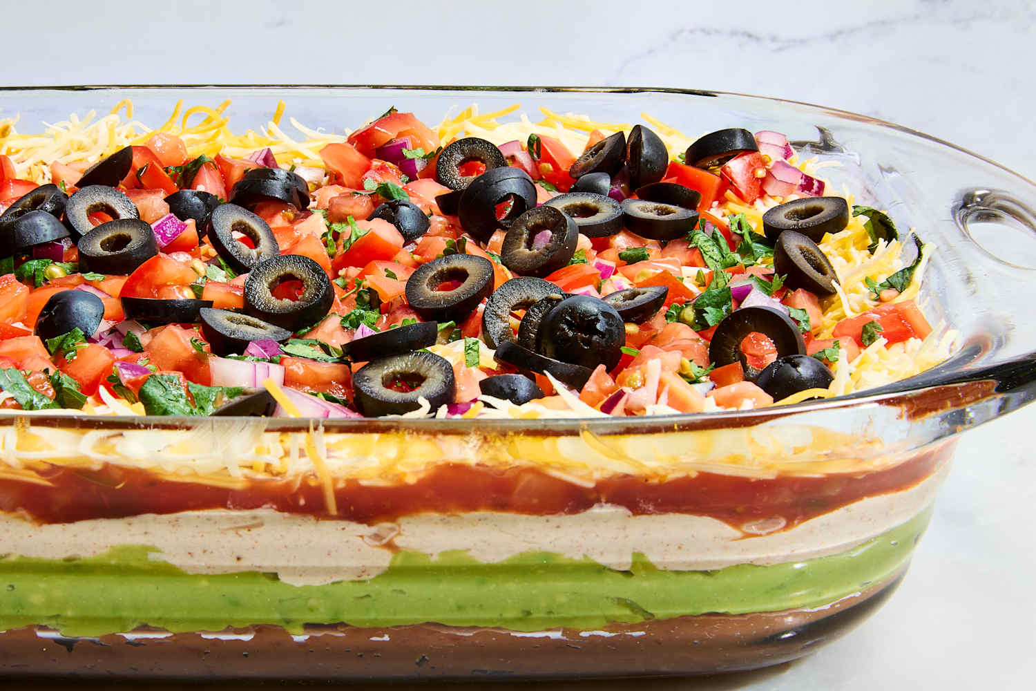 Close-up of Seven Layer Dip with a tortilla chip scooping through all the layers showing beans, guacamole, sour cream, cheese, and fresh toppings